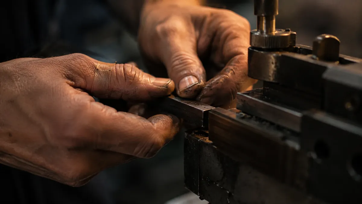 Zwei Hände setzen ein Werkstück in eine Maschine ein. Die Finger sind ölverschmiert, am Daumen klebt ein Pflaster.
